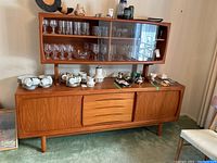 Front view of two-piece teak buffet hutch with glass upper cabinet and lower base displaying glassware (contents not included)