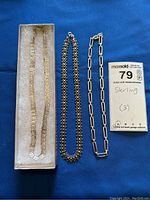 Photo showing three sterling silver necklaces arranged on blue fabric, one in a box with geometric square pattern design, a beaded strand, and rectangular link chain.