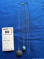 Three silver necklaces laid out on blue background including a pendant locket, a turquoise pendant, and a plain chain with broken clasp.
