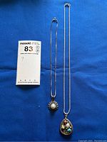 Two silver necklaces laid flat on blue fabric, showing full length and pendants.