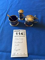 Set of sterling silver mustard pot, salt cellar, pepper shaker with small spoon on blue cloth, with note showing.