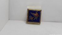 Square pillbox case on its side showing the blue enamel with bird design on lid
