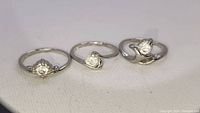 Three silver-tone rings with clear stones displayed side-by-side on a light surface.