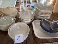Assorted baking dishes including ceramic pots and CorningWare dishes with clear glass lids displayed on table.