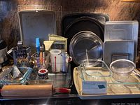 Photo showing a large metal baking sheet, round pizza pan, glass baking dishes, pizza cutter, rolling pin, and other utensils in a kitchen corner.