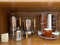 Photo showing three stainless steel coffee makers with gold handles alongside stacked brown ceramic espresso cups on saucers and clear glass cups on a white holder.