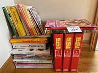 Cookbooks and magazines stacked on a wooden surface, titles mostly Italian culinary subjects.