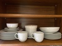 Wide shot showing assorted white embossed Denby plates, bowls, and cups on wooden shelf.