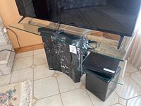Full view of the glass top TV stand with a TV and remote on it, accompanied by a small electronic device and subwoofer below the attached glass shelf.