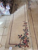Full view of the beige oriental carpet runner laid out on tiled floor, showing the floral border design along the edges and near the end, with a sofa partially visible.