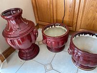 Set of three large planter pots in burgundy red ceramic placed on tiled floor near wooden cabinets. One tall urn-shaped pot and two round pots shown.