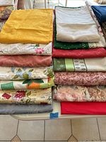 Photo showing two stacks of large folded tablecloths with various colors and patterns including mustard yellow, floral embroidery, red, green patterns, and newspaper print design.