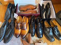 Overview of multiple men’s dress shoes arranged on floor including black and tan leather pairs, some in boxes, showing various styles such as slip-ons, lace-ups, and strap detail.