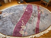 Full view of the round rug showing the gray background, maroon diagonal band, floral patterns, and fringe edges.