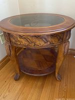 Front view of round wooden side table with glass insert and carved details on sides and legs.
