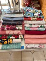 Image showing a large assortment of folded tablecloths with various colors and patterns, stacked on a dining table.