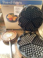 Copper metal hand press waffle iron with embossed maker stamp and extended handle next to electric waffle maker.