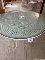 Full view of the round white iron patio table with glass top, showing the entire table and legs.