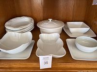 Photograph showing multiple white porcelain serving pieces including uniquely shaped bowls, covered casserole, square plates, and bowls arranged on a wooden shelf.