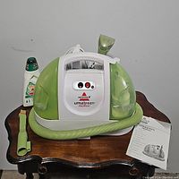 Front and top view of the Bissell LittleGreen ProHeat cleaner showing control panel, two tanks, hose, nozzle attachment, and user manual.