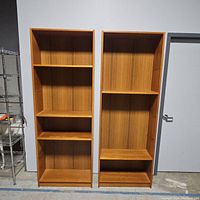 Two wooden bookcases side by side with multiple shelves, some shelves missing. Both have a natural wood finish and vertical grain lines.