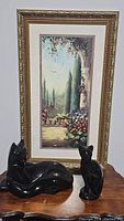 Photo showing both black cat statues placed on a wooden surface in front of a framed floral landscape painting