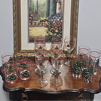 Full assortment of 13 Christmas-themed glasses: tumblers, stemless and stemmed glasses with tree and red accents on a table in front of painting.