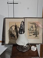 Two framed artworks showing city architecture placed behind a table lamp on a white surface and white background wall.