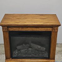 Front view of electric fireplace within oak finish wooden mantel showing faux logs and decorative columns.