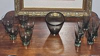Overview of all glass items on wood surface in front of a gold-framed painting: six tumblers, six shot glasses, one vase or bowl