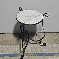 Side angle showing the marble top and wrought iron base with ornamental scroll design on a concrete floor.