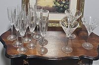 Both sets of stemware placed on a wooden table beneath a framed painting showing a group of champagne flutes and martini glasses.
