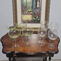 Eight pieces of stemware including three green-tinted champagne coupes and five clear wine glasses displayed on a small wooden table in front of a framed floral painting.
