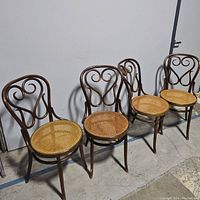Four vintage chairs with decorative curved wooden backs and woven rattan seats, positioned side by side against a white wall.