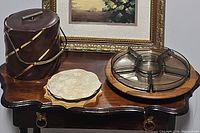 Photo showing entire lot on wooden table: brown leather and brass ice bucket, two vintage hot pads with floral design, and glass divided relish tray on wooden lazy susan.