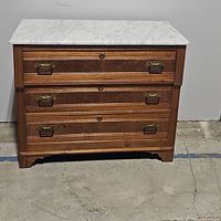 Front view of vintage wooden dresser with three drawers and white marble top, showing brass drawer handles and wood grain.
