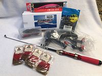 Fishing rod with red and black handle, box labeled Brocraft, package of artificial worms, and few packs of fishing lures and fly tying materials arranged on white background.