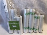 Photo showing 6 vacuum sealer rolls and bags, some with green stripe on surface, displayed on fabric background.