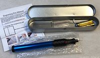 Engraving pen with blue and black body laid next to a silver metal case, instruction manual, and batteries on a white surface.
