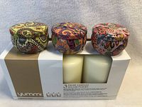 Photo showing box with three pillar candles in decorative tins with colorful patterned lids, labeled Yummi brand.
