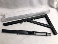 Photo showing two black folding brackets, one folded and one extended, along with mounting screws in bubble wrap.