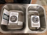 Stainless steel double sink shown with drain strainers and care instructions inside each basin.