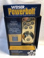 Front of the box showing the Weiser Powerbolt brand and electronic deadbolt lock visible inside, brass color