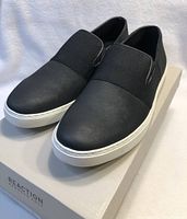 Angled side view showing the black upper and white sole of the Kenneth Cole loafers sitting on their box.