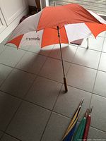 Orange and white 'allstream' branded golf umbrella open and standing on tiled floor showing wooden handle and metal shaft