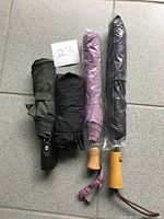 Four folded umbrellas placed side by side, two with wooden handles wrapped in plastic covers, two without, displayed on grey tiled floor.