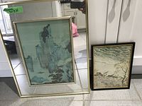 Photo of two Asian watercolor prints in frames; larger print showing mountainous landscape and smaller print with village scene under cloudy sky.