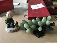 The photo shows the green jade grape cluster with agate carved leaves alongside the porcelain monk figurine with accompanying small chicken and gourd on white textured base.