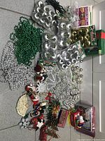 Photo showing assortment of Christmas hanging ornaments, bead strings, and boxed ornaments on tiled floor