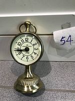Round brass-finish tabletop clock with clear view of clock face and stand.
